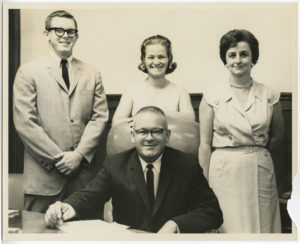 John Tate, Patricia Howell Tate, Lula "Puggie" Howell, and Max Howell pose for a group portrait in an office. Max Howell is sitting in his office chair while the other family members are standing behind him.