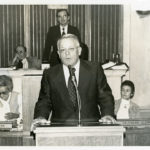 Max Howell speaking at podium in the Arkansas State Senate chambers. He is speaking to Senate colleagues. Two women are sitting behind Howell and two men, with one standing, are at the dais.