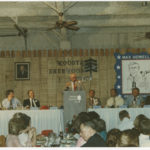 Max Howell speaks at the ninth annual Max Howell Roast of state Senator Ben Allen. Howell is standing at the podium with five men seated next to him. In the foreground, several attendees are seated.