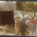 Huckaby stands to the side of the movie set, in the outdoors, with a truck parked on the street, and other equipment placed on the sidewalk.