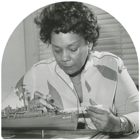 Black and white photograph of Raye Montague sitting at a desk looking at a model ship.