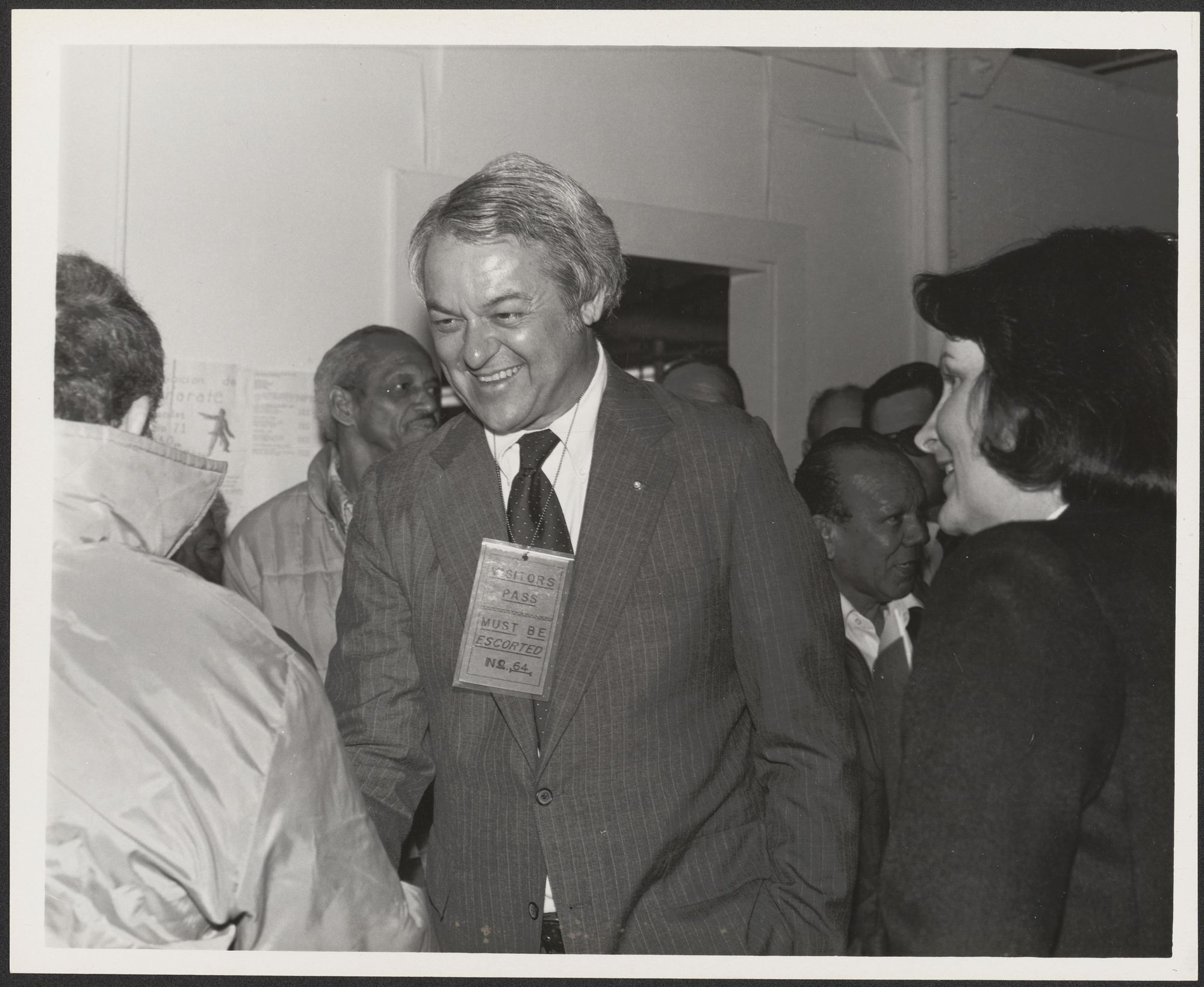 A black and white photograph of Governor Frank White shaking hands at Fort Chaffee. He has a visitors pass around his neck.