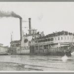 Black and white photograph shows the overloaded steamboat Sultana on the Mississippi River. Black smoke pours out of the left smokestack and the upper and lower decks are filled with Union soldiers in dark military uniforms. The steamboat is pointed to the right in the image, downriver, with the word SULTANA in large script in the middle of the steamboat. There is a smaller barge to the rear in the bottom left of the photograph.