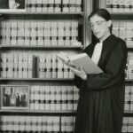 Black and white photograph of United States Supreme Court Justice Ruth Bader Ginsburg standing on a stool reading a law book in front of a bookcase