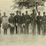 Onlookers stand on the steps of a building while 13 members of the 57th United States Colored Infantry, an African American unit in the Civil War, pose in uniform for the photographer. The three men on the left carry a bugle, large drum, and snare drum. Other men hold weapons. The tallest man in the center carries the American flag.