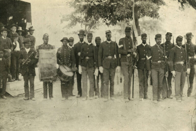 Onlookers stand on the steps of a building while 13 members of the 57th United States Colored Infantry, an African American unit in the Civil War, pose in uniform for the photographer. The three men on the left carry a bugle, large drum, and snare drum. Other men hold weapons. The tallest man in the center carries the American flag.