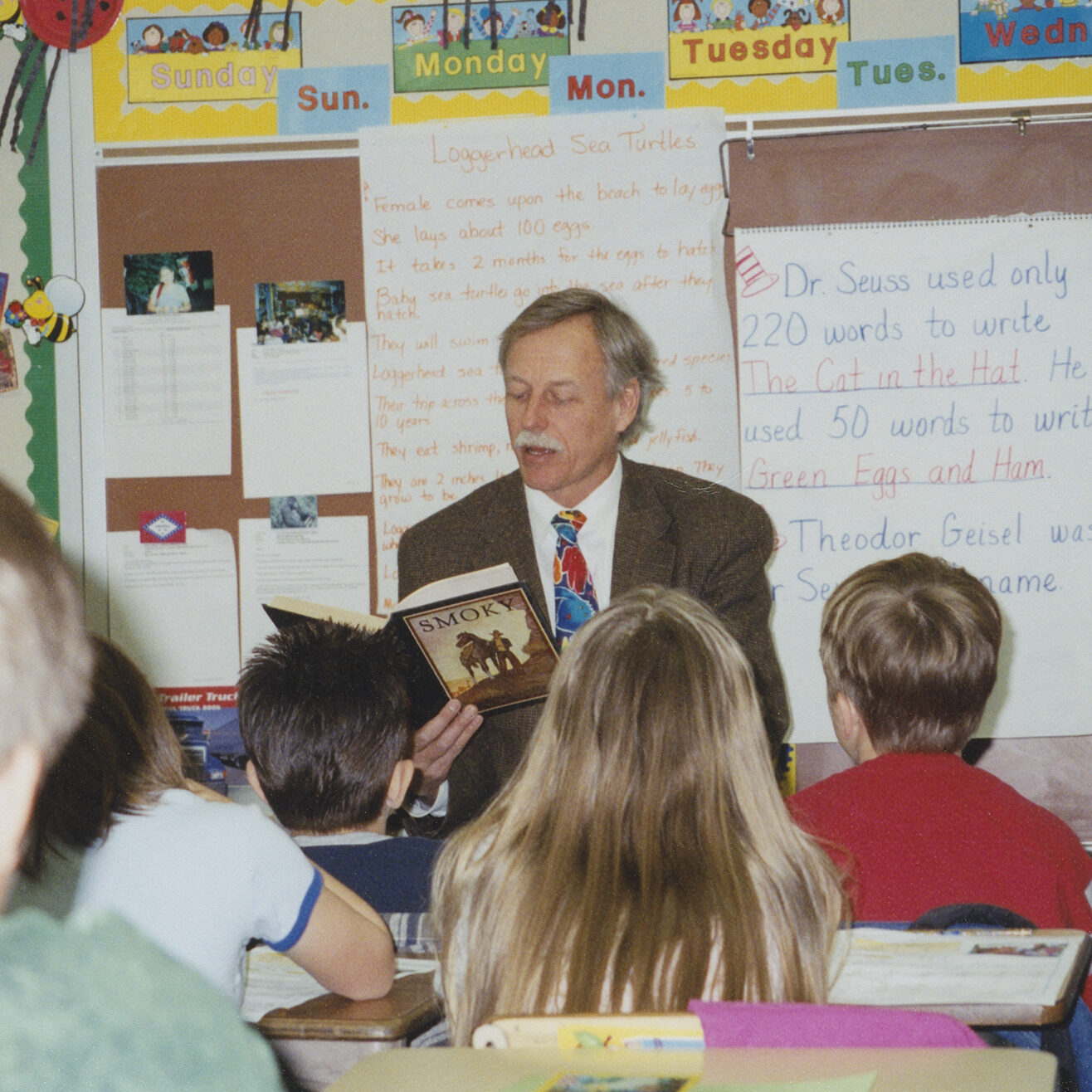 Color photograph of Snyder sitting at the front of a classroom and reading "Smoky the Cow Horse" to a group of students