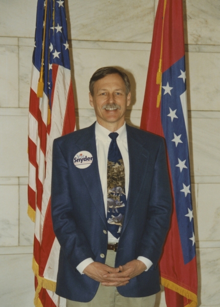 Snyder wearing his campaign button and standing in front of the American and Arkansas flag, 1996