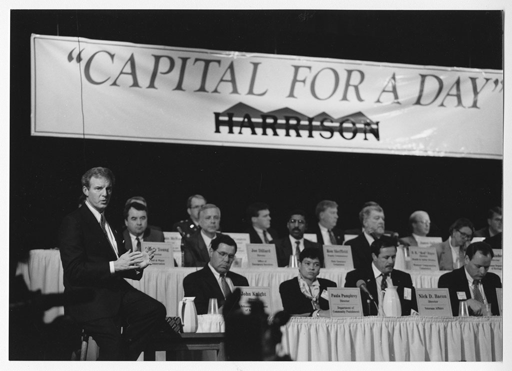 Jim Guy Tucker in front of cabinet and Capital for a Day sign in Harrison, Arkansas