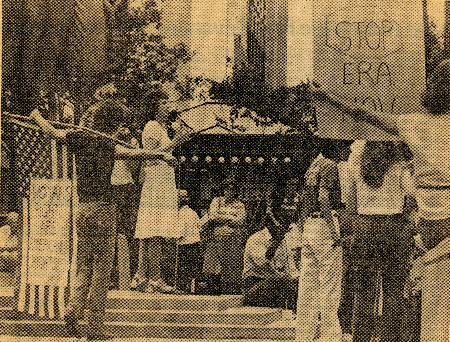 Newspaper photograph of a rally for the ERA. One woman gives a speech. Supporters hold posters that read, "Women's rights are American Rights," and a protester holds a sign that reads, "Stop ERA now.